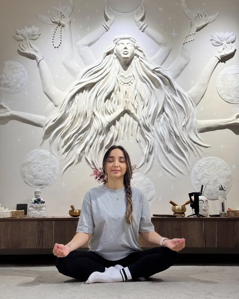 Gunel sitting in a meditation pose with her eyes closed in front of a white relief sculpture